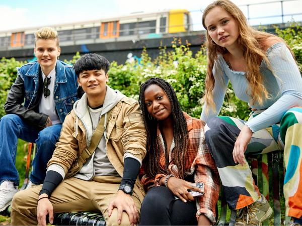 Four young people sat on a wall looking towards you and smiling at the camera